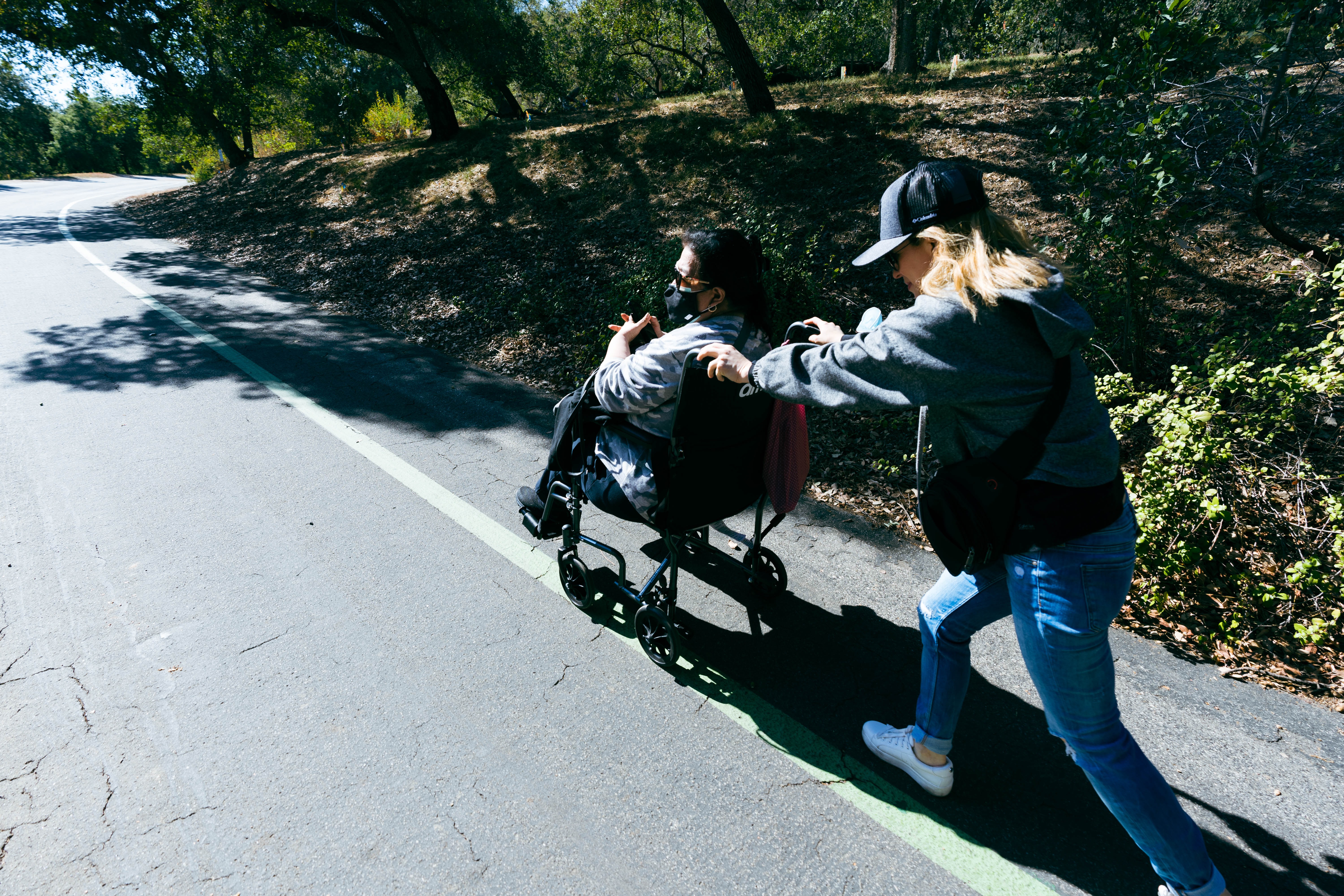 A woman pushing another woman in a wheelchair up a paved hill